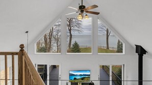 A spacious room with a sloped ceiling, a ceiling fan, and multiple windows offering views of a lake and trees. - HOT TUB | Lake Front | Lavender Home (Burt)