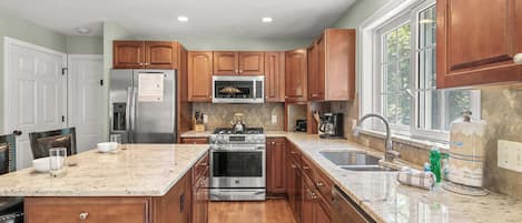 A kitchen with wooden cabinets, stainless steel appliances, a large window, and a marble countertop.