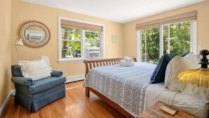 A bedroom with a bed, a chair, a lamp, and large windows. - Close to Bike Path 1 Mile to Beaches! (South Chatham)