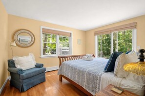 A bedroom with a bed, a chair, a lamp, and large windows. - Close to Bike Path 1 Mile to Beaches! (South Chatham)