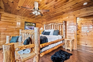 A wooden cabin bedroom with a bed, a chair, a small table, and a ceiling fan. - Vet-Owned, Pet Friendly Pool Cabin with New Hot Tub & Amazing Views! (Pigeon Forge)