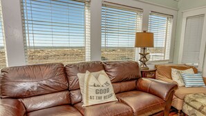 Ein braunes Ledersofa mit einem Kissen, auf dem 'LIL'S GOOD AT THE BEACH' steht, in einem Wohnzimmer mit großen Fenstern und einer Lampe.