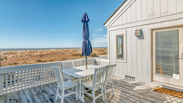 Eine Holzterrasse mit einem weißen Tisch und Stühlen, einem blauen Sonnenschirm und Blick auf den Strand.