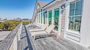 A wooden deck with two lounge chairs and a small table, overlooking a beach. - Reverse floor plan home with ocean views, updated kitchen and bathrooms! (Bald Head Island)