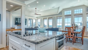 A spacious kitchen with a large island, wooden dining table, and chairs, overlooking a scenic view. - Reverse floor plan home with ocean views, updated kitchen and bathrooms! (Bald Head Island)