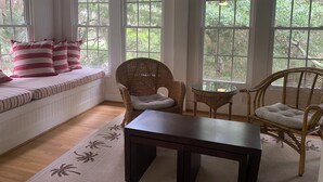Una habitación acogedora con un banco empotrado, sillas de mimbre, una mesita pequeña y ventanas grandes con vistas al verdor.