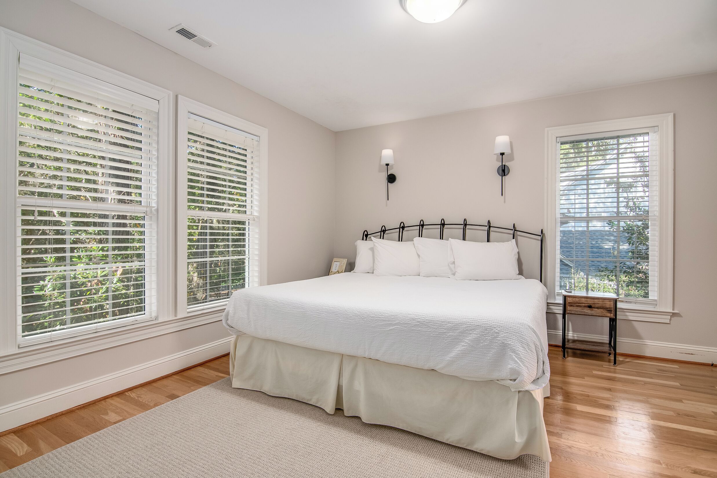 A bedroom with a large bed, white bedding, two wall-mounted lamps, and large windows with blinds.