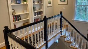 Una escalera con barandilla negra y balaustres blancos que conduce a una plataforma con estanterías y obras de arte enmarcadas.