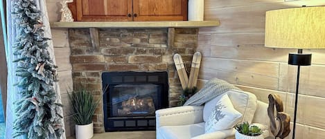 A cozy living room with a fireplace, a white armchair, a small side table, and a Christmas tree.