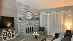 Ein Wohnzimmer mit Backsteinwand, Kamin, Fernseher, einer Couch und einem Couchtisch mit Blumen.