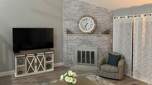 Ein Wohnzimmer mit Backsteinwand, Flachbildfernseher, einer Couch, einem Couchtisch mit Blumen und einem Kamin.