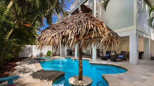 A pool area with a thatched umbrella, lounge chairs, and a palm tree. - Fish Stix: Elevator, 4 Houses to Beach, Pool by Anna Maria Life Vacation Rentals (Holmes Beach)
