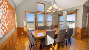 Comedor con mesa y sillas de madera, ventanas amplias con vista a la playa y un candelabro.