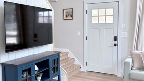 Una sala de estar con un televisor de pantalla plana, un mueble azul, una escalera, una puerta blanca y un sofá de color claro.