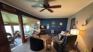 Un salon chaleureux avec un ventilateur de plafond, un canapé vert, un fauteuil en cuir noir et une table basse.