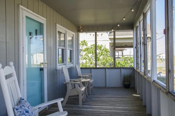 A covered porch with wooden chairs and a bench, large windows, and a view of greenery. - Adorable Beach Box, Very short walk to the beach Access! (Kill Devil Hills)