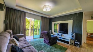 A modern living room with a large flat-screen TV, a sofa, a chair, and a balcony with curtains.