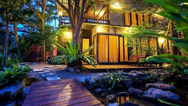 Una casa moderna con terraza de madera, rodeada de plantas tropicales y un pequeño estanque.