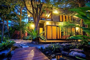 Uma casa moderna com deck de madeira, cercada por plantas tropicais e um pequeno lago.
