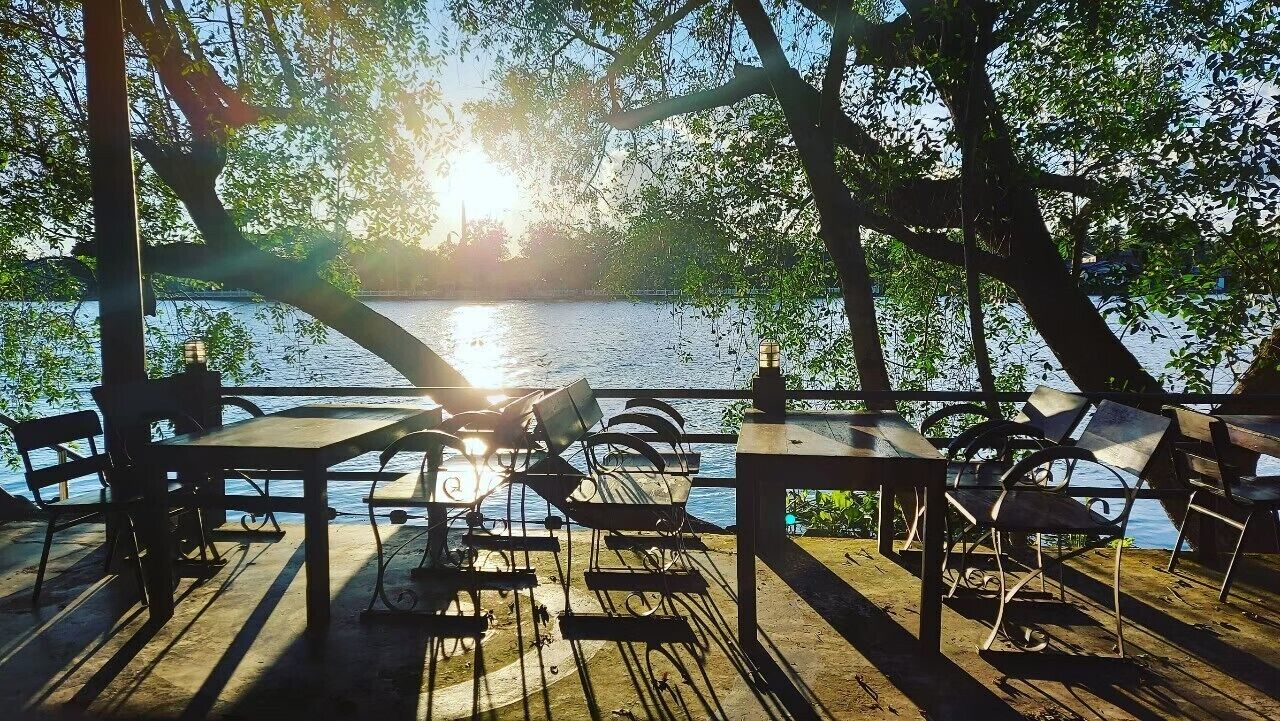 Terraza de madera con mesas y sillas, con vista a un cuerpo de agua, donde la luz del sol se filtra entre los árboles.