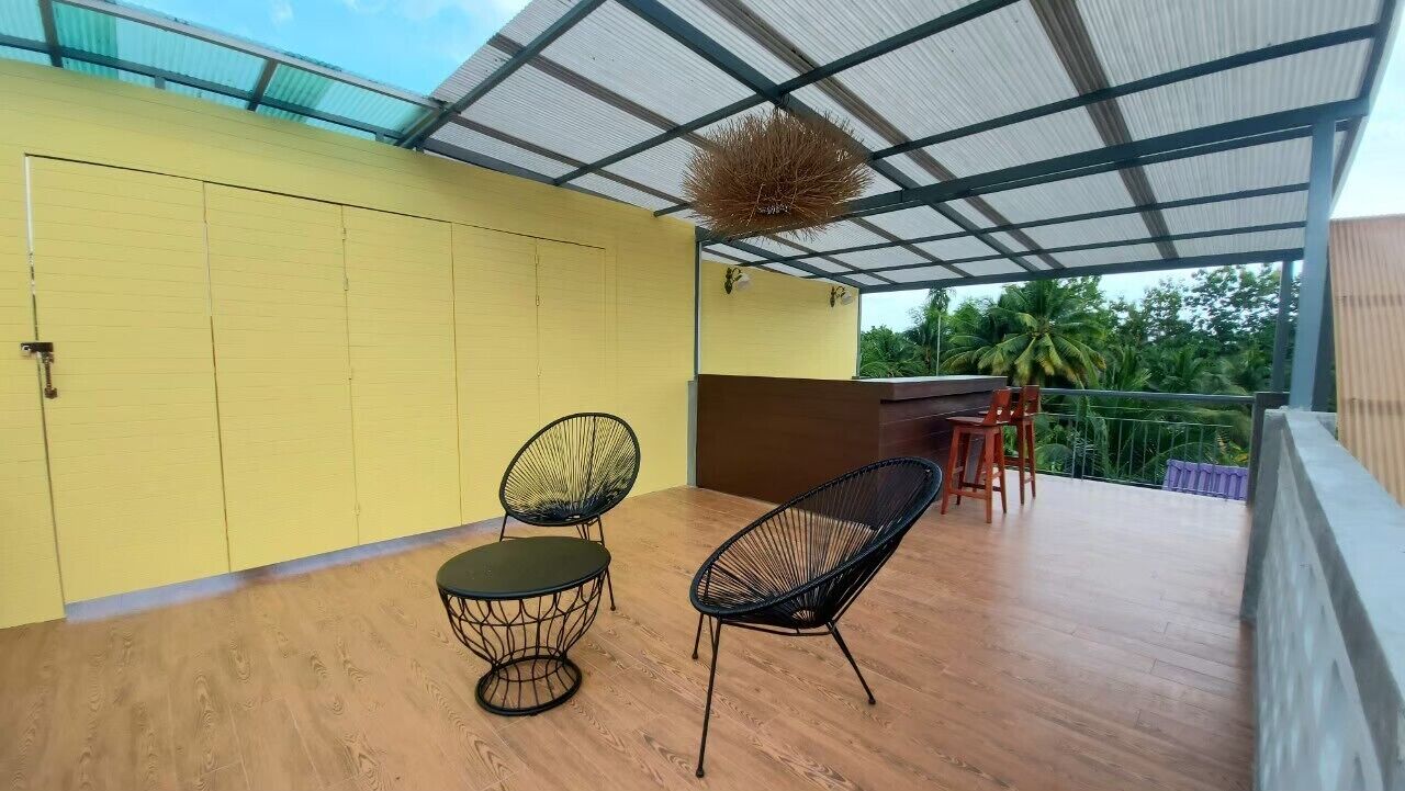 A covered outdoor area with a yellow wall, two black wireframe chairs, a small round table, and a wooden deck.