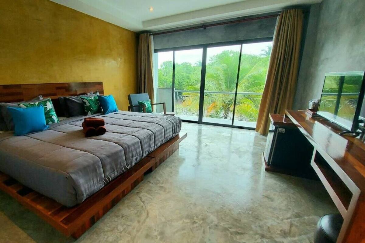 Une chambre spacieuse avec un grand lit, une tête de lit en bois, un bureau et une vue sur la verdure grâce à de grandes fenêtres.