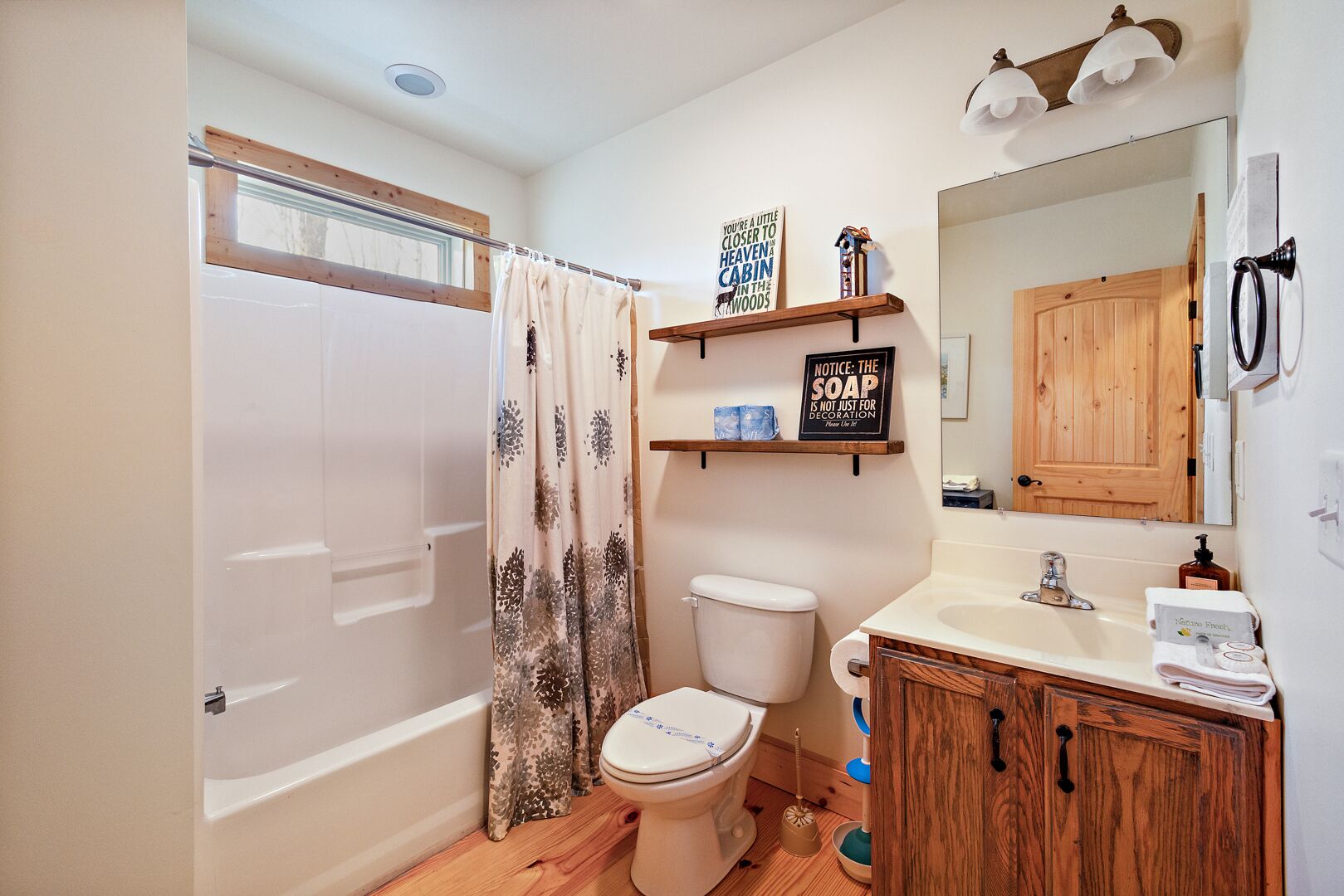 Een badkamer met toilet, douchegordijn, houten wastafel en een spiegel. Boven het toilet staan twee planken met decoratieve objecten.