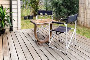 A wooden deck with a metal table and two chairs, a glass of wine on the table, and potted plants around.