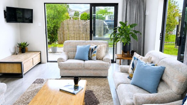 A living room with two sofas, a coffee table, a TV, and a sliding glass door leading to a balcony. - 3BR Home • Parking • Sleeps 6 • Near Transport (Panmure)