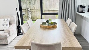 A modern dining area with a wooden table, white chairs, and a small potted plant centerpiece. - 3BR Home • Parking • Sleeps 6 • Near Transport (Panmure)