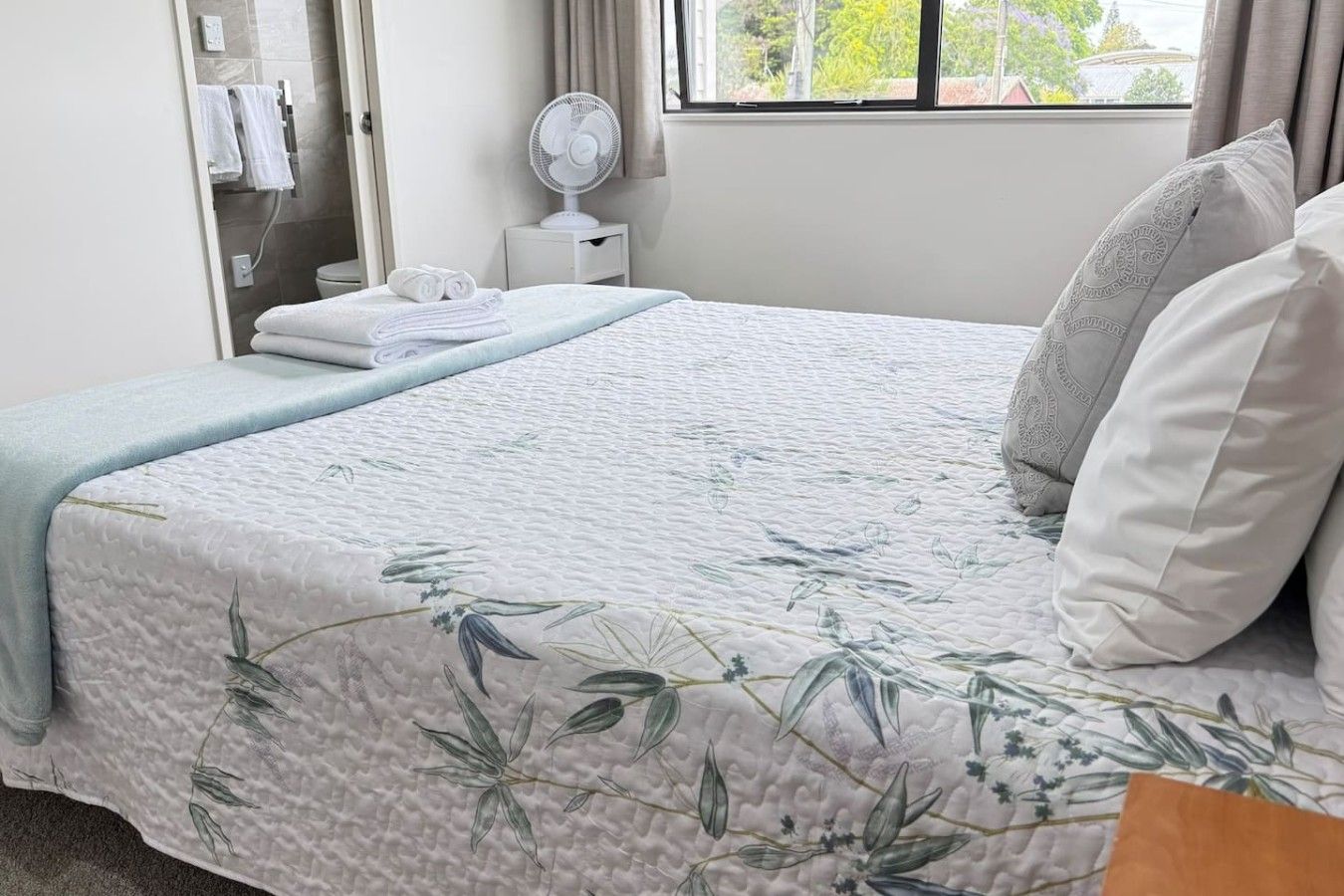 A neatly made bed with a floral-patterned bedspread, a bedside table with a fan, and a window with a view of greenery.