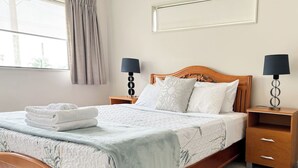 A neatly made bed with a white comforter and pillows, a bedside table with a lamp, and a window with curtains. - 3BR Home • Parking • Sleeps 6 • Near Transport (Panmure)