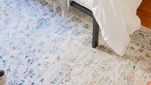 A bed with a white sheet and a beige blanket, a patterned rug, and a plain wall. - Vintage Escape 1BR Two Blocks Away From Beach (Chicago)
