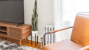 A living room with a TV on a wooden stand, a potted plant, a radiator, and a brown leather chair. - Vintage Escape 1BR Two Blocks Away From Beach (Chicago)