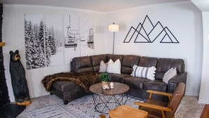 Un salon avec un canapé d’angle en cuir marron, une table basse et un tapis à motifs. Un mur est décoré d’œuvres d’art encadrées et il y a une lampe.