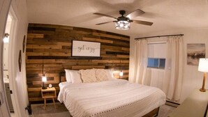 Une chambre avec un lambris en bois, un ventilateur de plafond, un lit avec une literie blanche, une table de chevet avec une lampe et une fenêtre avec des rideaux.
