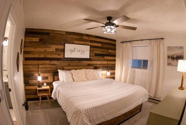 Un dormitorio con panel de madera en la pared, ventilador de techo, cama con ropa blanca, mesita de noche con lámpara y ventana con cortinas.