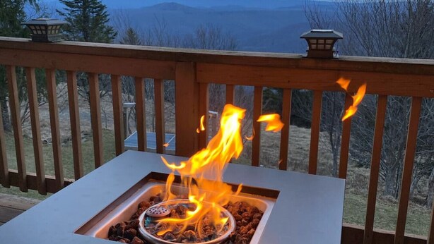 Un foyer extérieur carré, doté d’une grille en métal et de bois qui brûle, entouré d’une terrasse en bois avec des garde-corps.