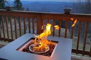Fuego al aire libre cuadrado con rejilla de metal y leña ardiendo, rodeado por una terraza de madera con barandillas.