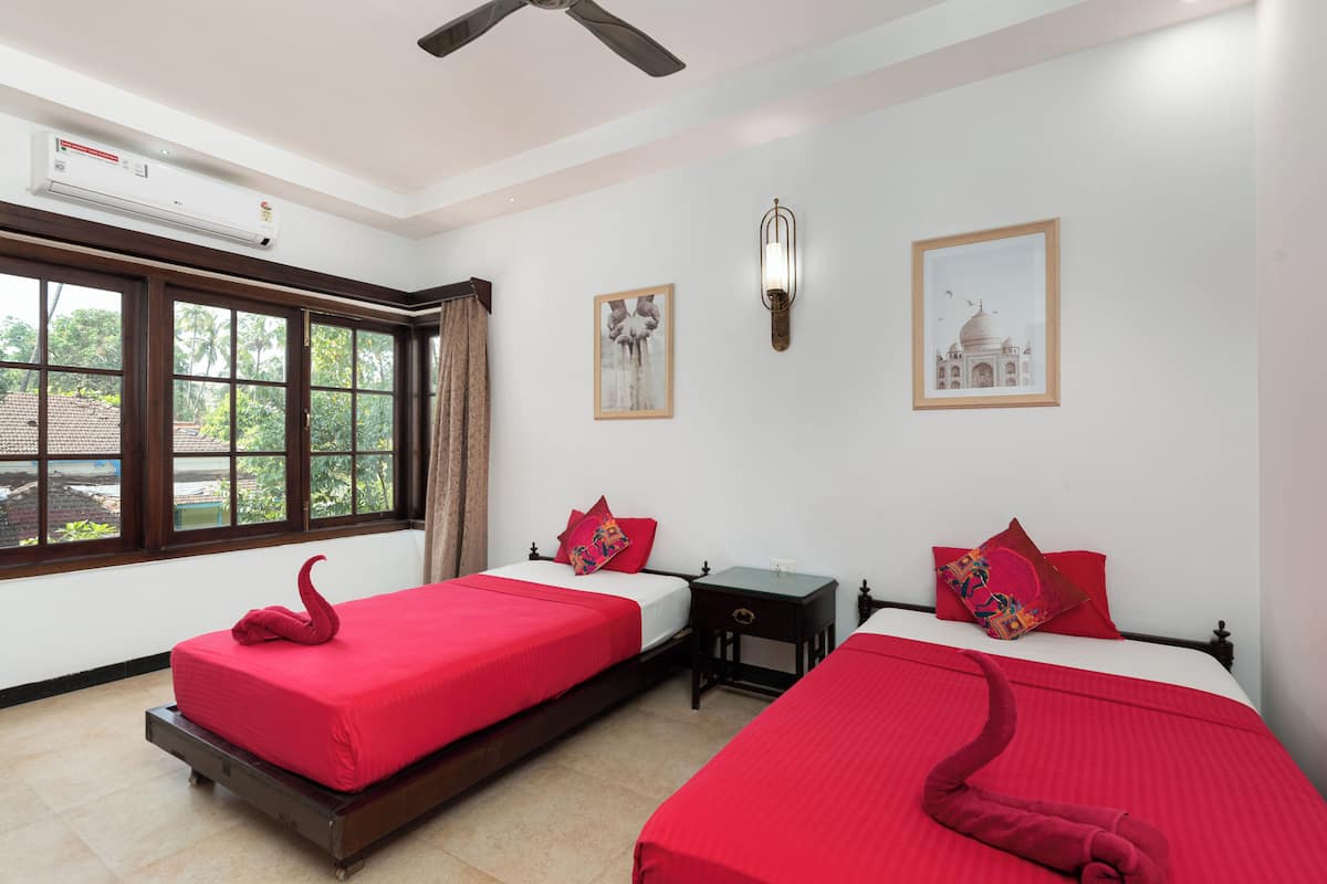A hotel room with two beds, a ceiling fan, an air conditioning unit, a window with curtains, and two wall-mounted light fixtures.