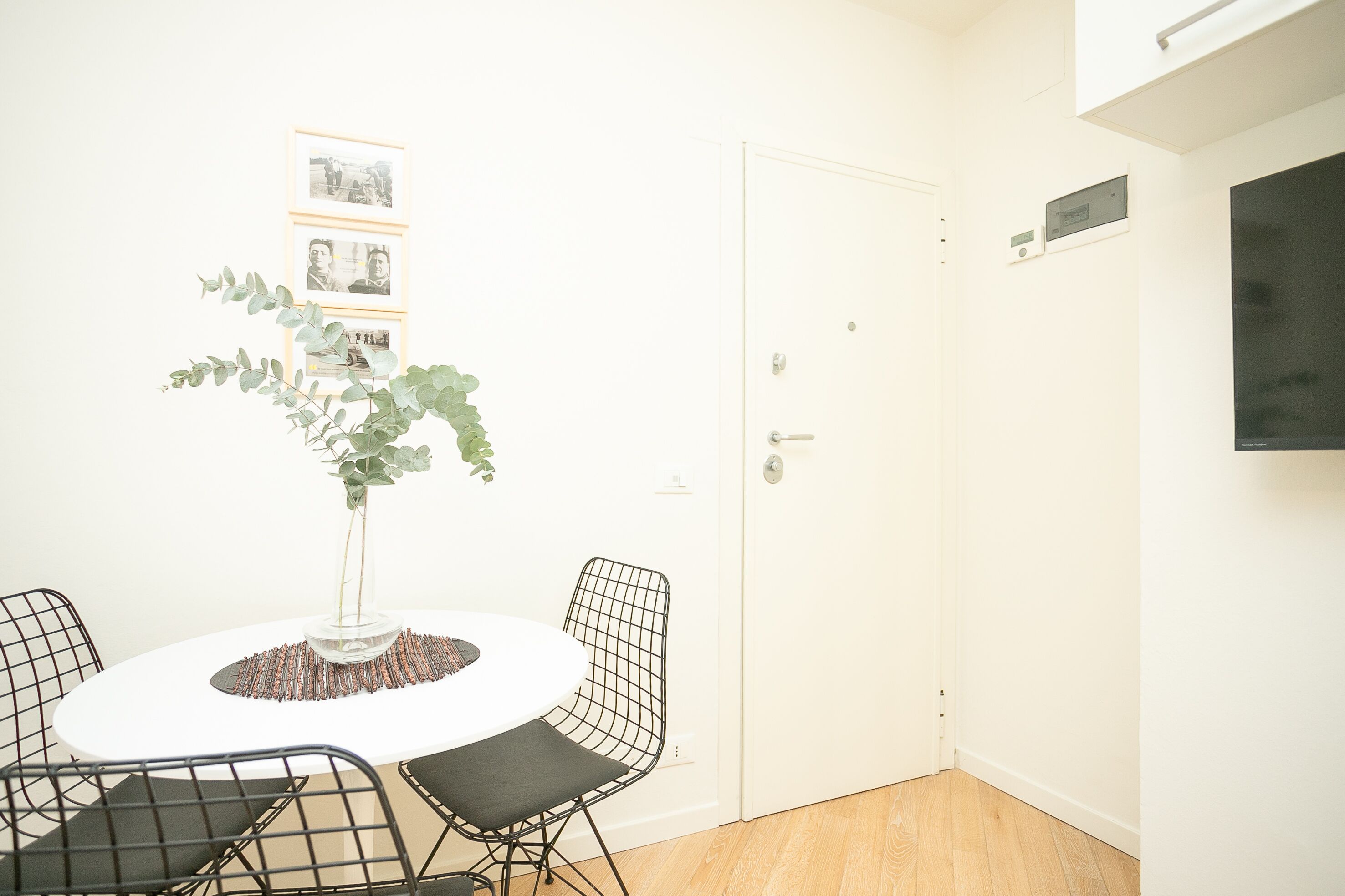 A room with a round table, four wireframe chairs, a potted plant, and framed pictures on the wall.