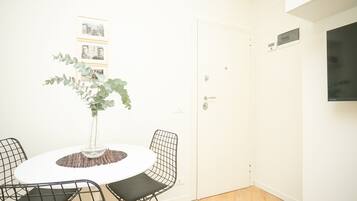 A room with a round table, four wireframe chairs, a potted plant, and framed pictures on the wall.
