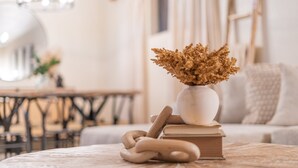 Una mesa redonda de madera con un jarrón blanco que contiene flores secas, una pila de libros y un par de cucharas de madera.