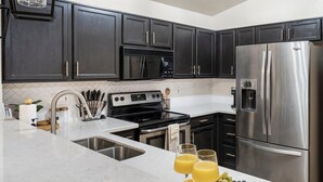 Cocina moderna con armarios oscuros, electrodomésticos de acero inoxidable, encimera blanca y una bandeja de frutas.