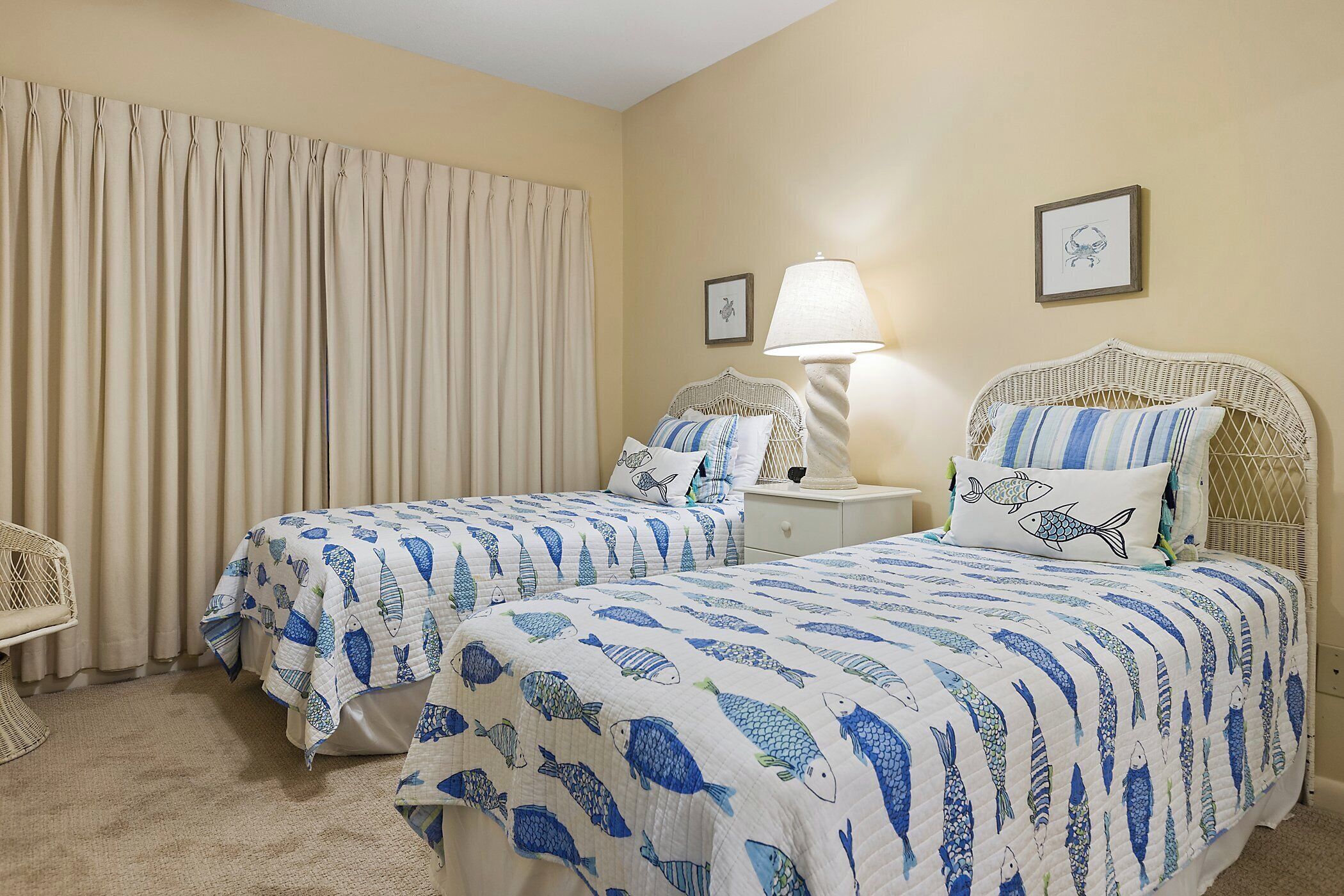 A room with two beds, each with a blue and white fish-patterned bedspread, a wicker chair, a lamp, and a framed picture on the wall.