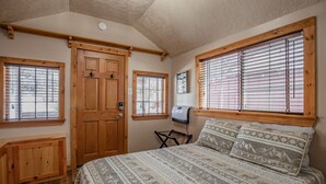 A bedroom with a wooden door, two windows with blinds, a bed with patterned bedding, and a small table with a heater.