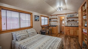 A bedroom with a bed, two windows, a kitchenette, and wooden cabinets.
