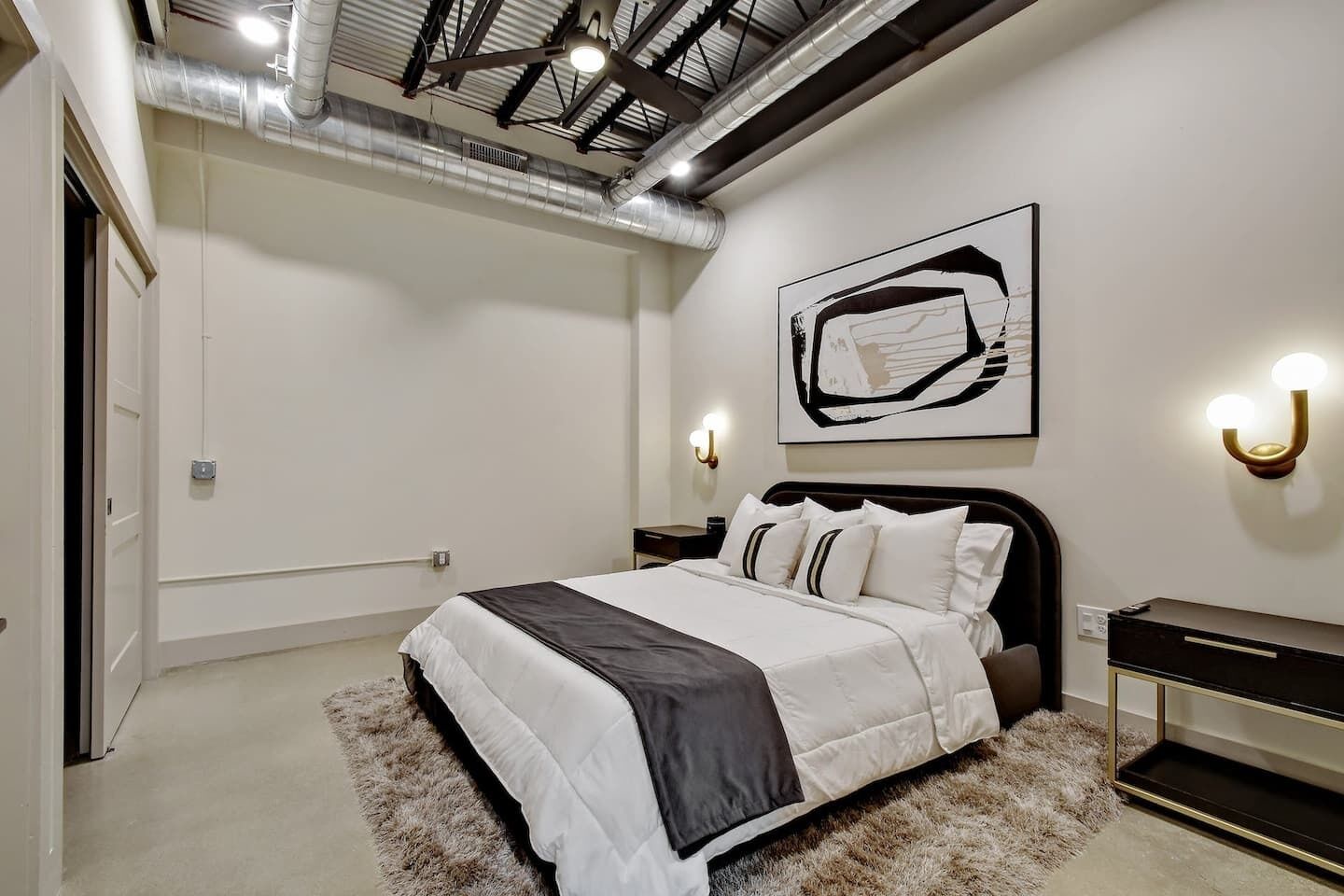 Een moderne slaapkamer met een groot bed, een hoofdbord, nachtkastjes, een kunstwerk aan de muur en een plafond met blootgelegde leidingen.