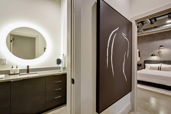 Un baño moderno con un espejo redondo, un tocador con un mueble oscuro y una obra de arte grande con el rostro de una mujer en la pared.