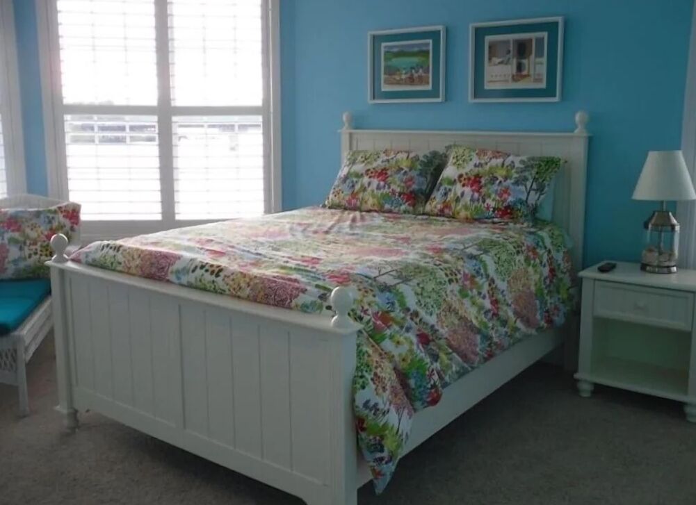 A bedroom with a white bed, floral bedding, a nightstand with a lamp, and two framed pictures on the wall.